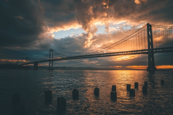 Image shows the Golden Gate Bridge with a background of a cloudy sky, the sun peaking just above the horizo and reflecting on the water.